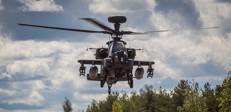 A military attack helicopter flying low above a forested area under a partly cloudy sky.