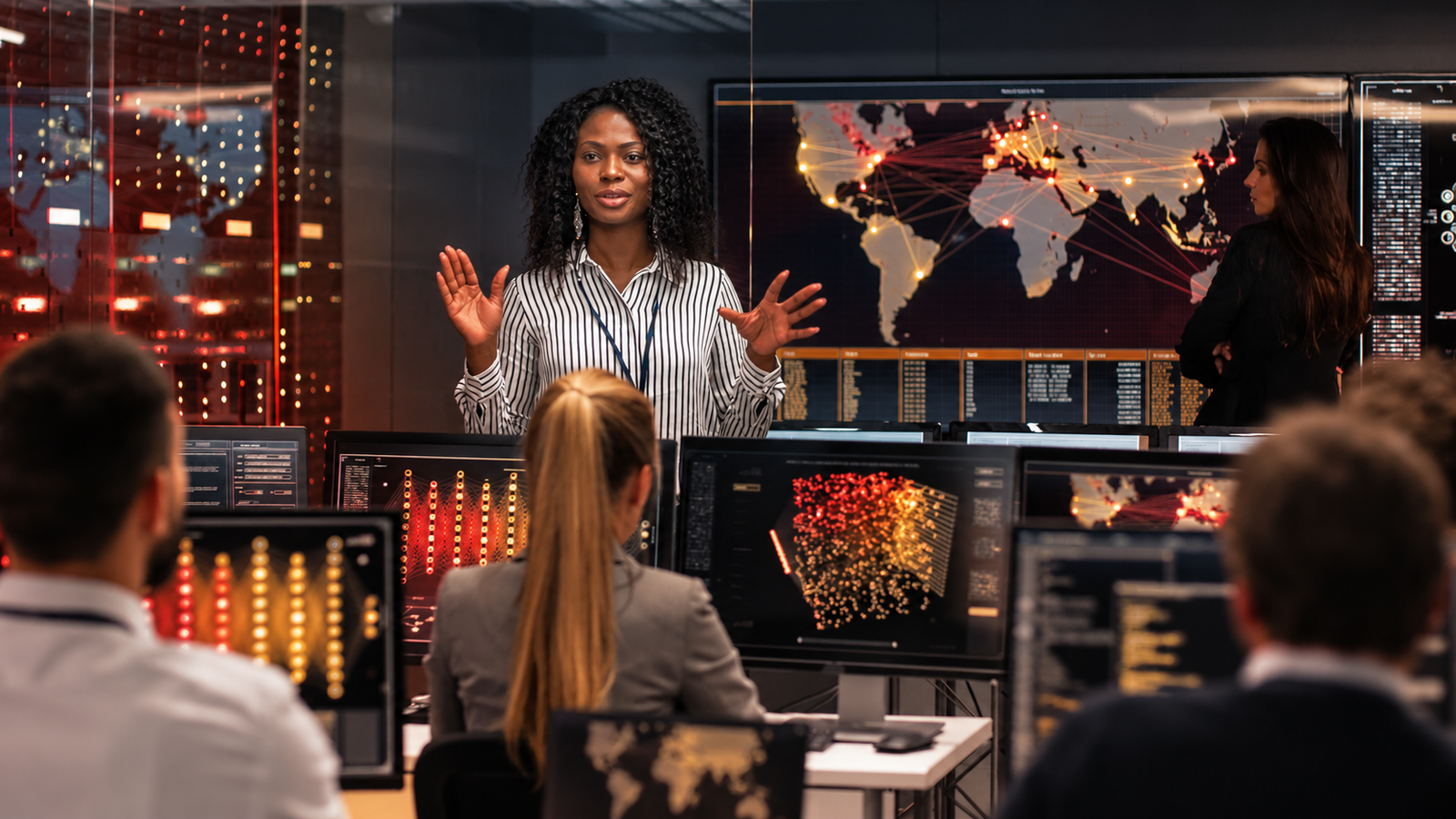 Presenter addressing a team in a dark control room with world map and data displays on screens.