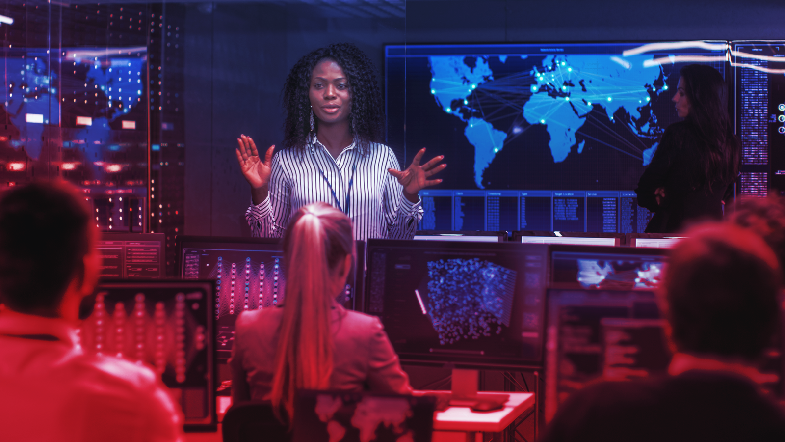 Presenter addressing a team in a dark control room with world map and data displays on screens.