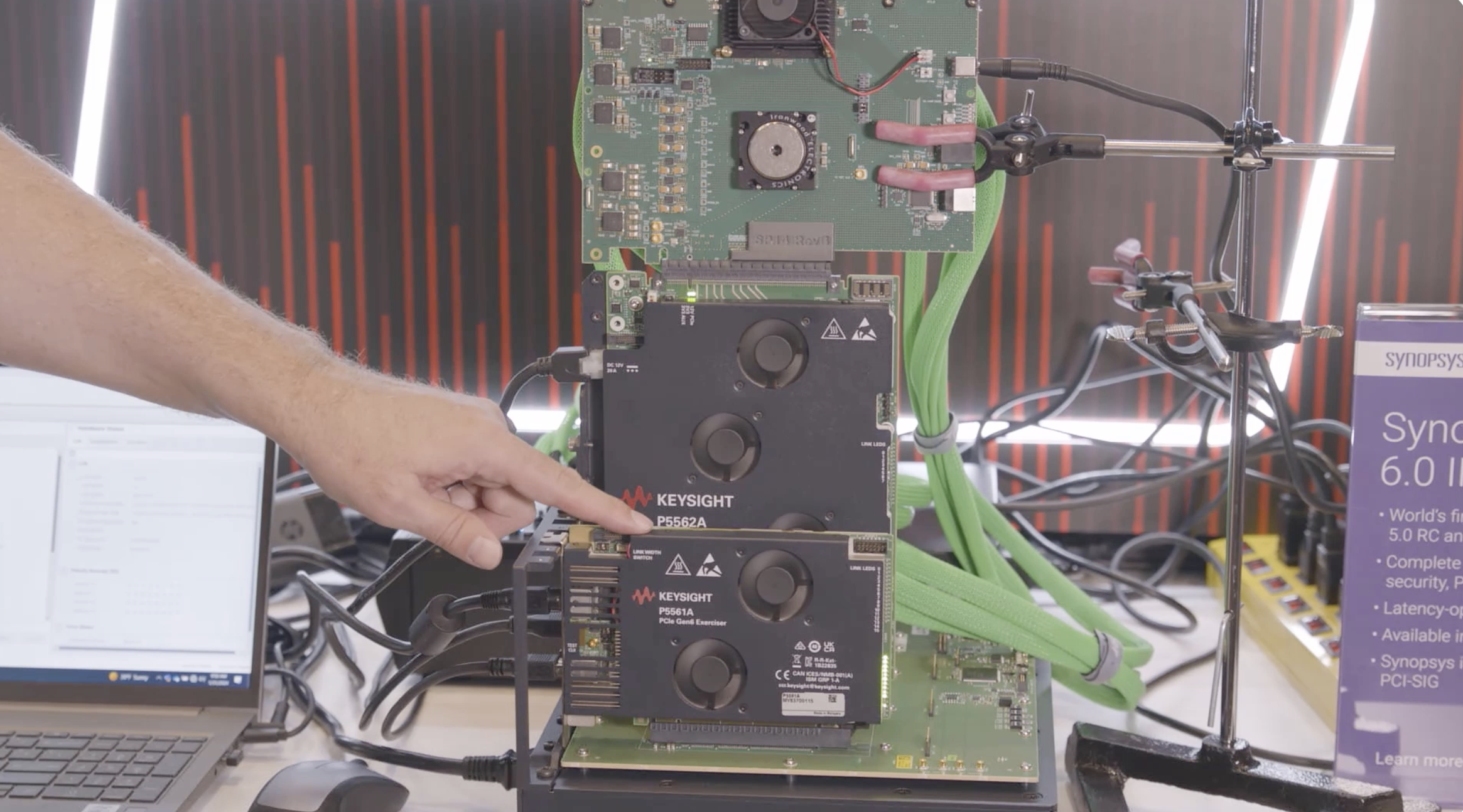 Eine Person zeigt auf einen Keysight P5561A PCIe Card Exerciser, der Teil eines Multi-Board-Testaufbaus ist, der das Keysight P5562A Base Board und verschiedene angeschlossene grüne Kabel umfasst. Das System ist vertikal montiert und dient der Validierung und dem Testen von PCI Express.